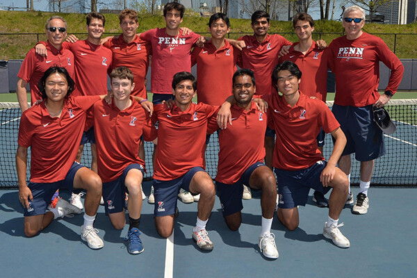On a tennis court, coaches and players on the men's tennis team pose sitting and standing with their arms across each other's shoulders.