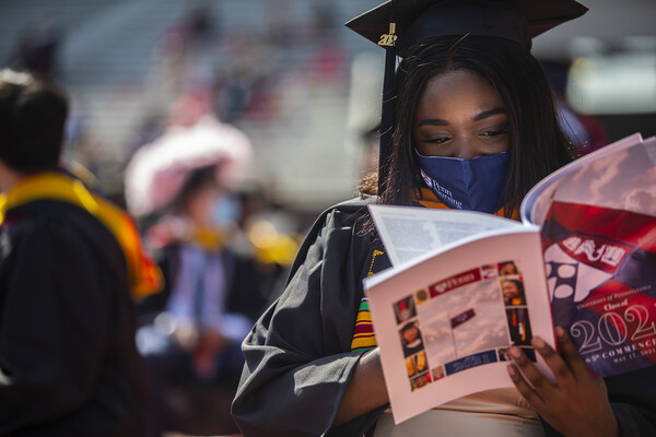 A masked person in graduation robes and cap looks at a program.