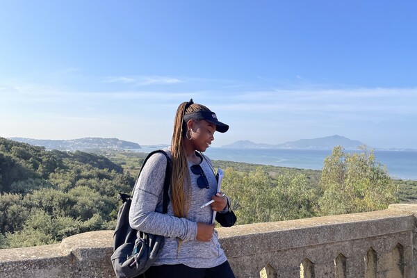 A woman wearing a visor and backpack holds a notebook in front of a stone wall with mountain views in the background