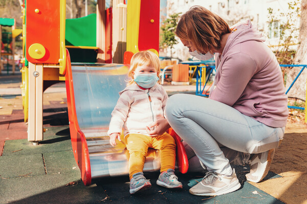 An adult wearing a mask squatting next to a child wearing a mask at the end of a slide on an outdoor playground.