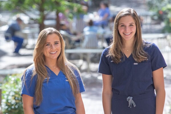 Two smiling women stand in blue medical scrubs on a sunny day.