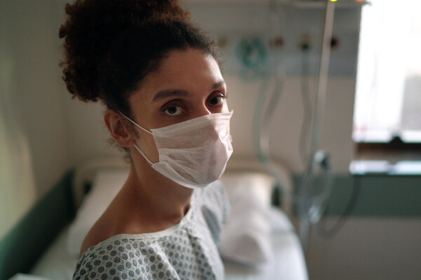 A transgender patient wearing a mask and hospital gown on a hospital bed.