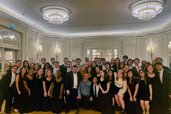 Glee Club members in formalwear gathered together in ballroom under crystal chandeliers