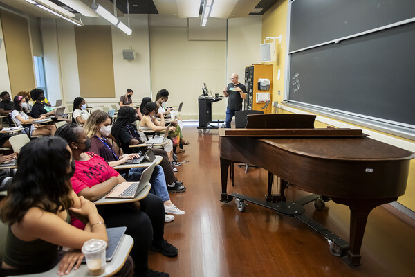 Timothy Rommen next to a piano and a podium teaching a class full of students.