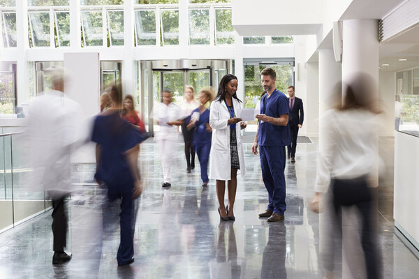 Scene of a hospital lobby. Most people are blurry, but two are clear, a person in a lab coat talking to a person in scrubs. 