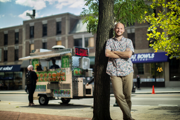 Max Johnson Dugan leans against a tree in front of a halal food cart on the street.