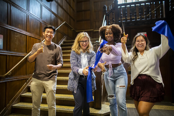 President Liz Magill cuts the ceremonial ribbon with three students. A wood-paneled staircase is the backdrop 