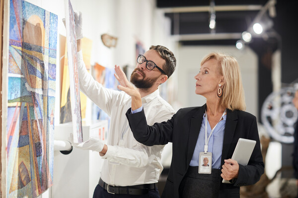 Two people standing in front of a wall of art. One of them is holding up a second piece of art in gloved hands. The other gestures toward the art, holding a computer or clipboard in the other hand. 