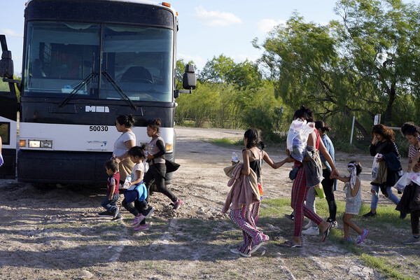 A group of people carrying plastic bags cross a dirt road towards a bus