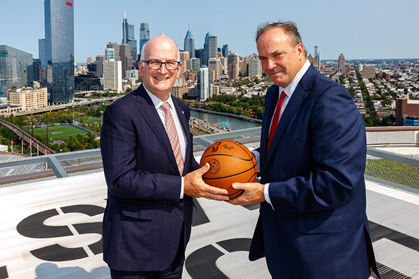 Two men stand outside against Philadelphia backdrop both holding a basketball between them
