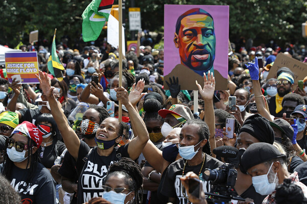 A crowd of people outdoors, some wearing masks, some with arms raised, some holding signs that read "Justice for George Floyd" or with a painting of George Floyd's face.
