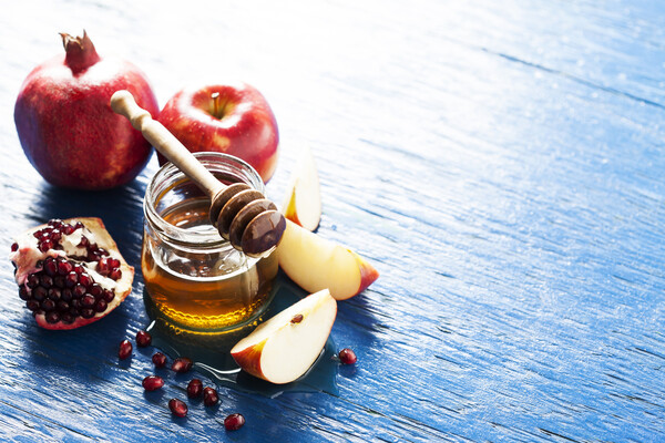 A cut apple and pomegranate surround a honey jar with a wooden honey stick on top. An uncut apple and pomegranate are in the background.