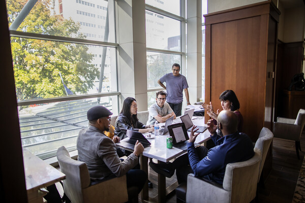 five people sitting around table with laptops open and one person standing