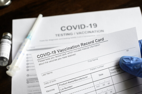 A gloved hand holds a COVID-19 Vaccination Record card. Blurred in the background are a closed needle, a vial of medication, and a sheet of paper that reads "COVID-19 Testing/Vaccination." 