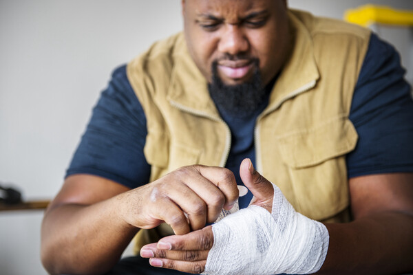 Person with a goatee and moustache wearing a tee-shirt and vest, the hand wrapped up in gauze as if it was injured. The person has a pained expression.