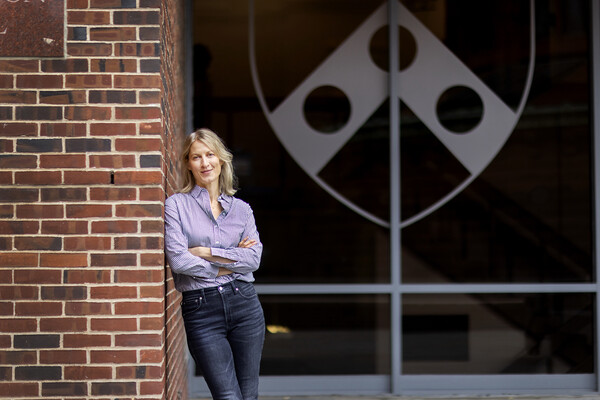 Economics professor Anne Duchene leans up against a red brick wall with her arms crossed.