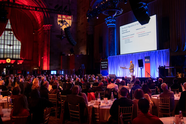 Lance Freeman, at a podium on a stage, speaks to a crowd with a presentation displayed in the background