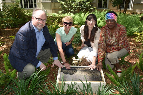 Four people kneel outside in front of a memoerial plaque, each person with one hand touching it. The words "African Burial Ground ca. 1750-1800" are visible.
