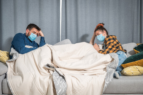 two young people wearing lie under a blanket on a couch, looking sick