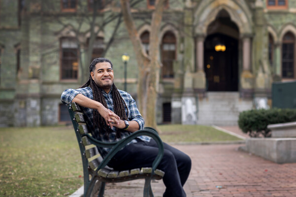 Daniel Morales-Armstrong sits on a park bench in front of Penn's College Hall