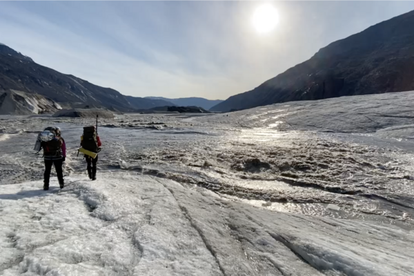 Two scientists walk on glacier ice near a river and mountains