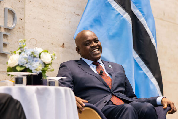 The president of Botswana smiles as he sits on a stage in front of the flag of his nation next to a bouquet of light blue and white flowers