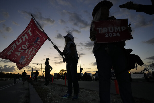 Trump supporters hold Trump 2024 flags and campaign signs in shadow as the sun sets.