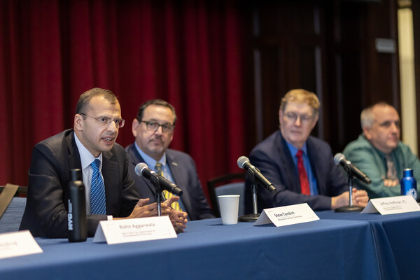 Four panelists sit at table listening while one speaks into microphone