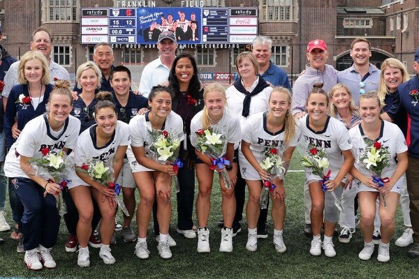 Seven seniors on the women's lacrosse team pose on Franklin Field with flowers after the Cornell game. Coaches and family members stand behind them.
