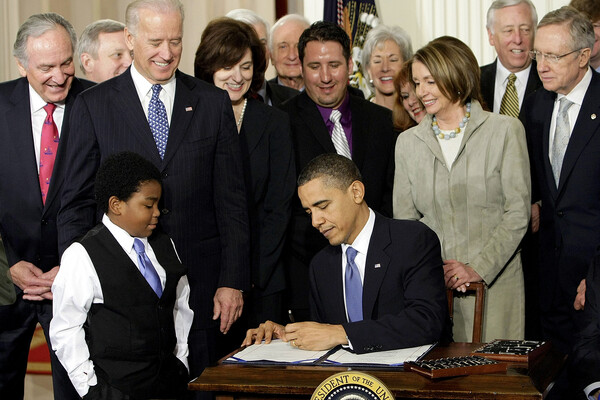 President Barack Obama signs the Affordable Care Act surrounded by lawmakers and a young child standing by the table.