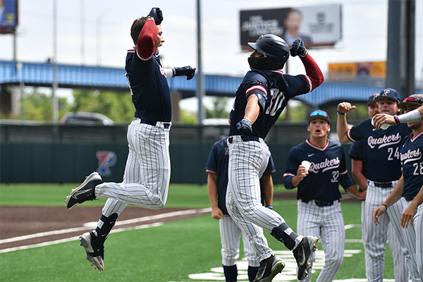 Wearing their blue Penn jerseys, two baseball players jump in the air and prepare to slap arms in celebration.