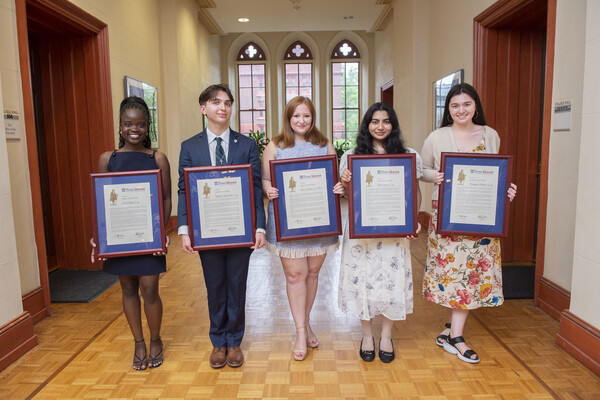 Five students holding framed certificates stand in a hallway