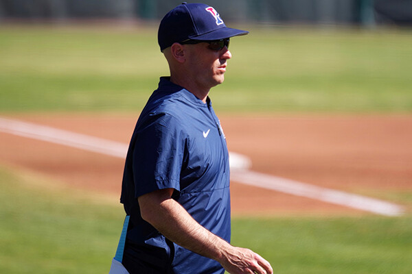Manager John Yurkow, wearing a blue Penn hat and shirt and black sunglasses, observes play on the field from the sidelines.
