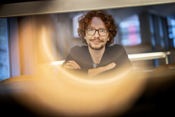 Man with red curly hair and glasses looks into the camera, which is giving a half-moon glow on the lower right side of the image.