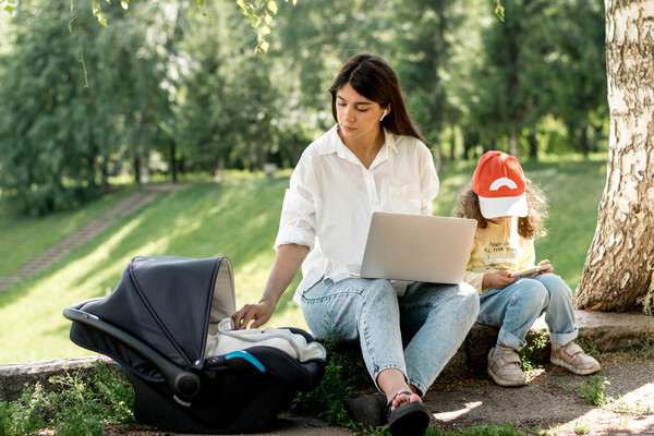 A childcare worker outside in a park with a toddler and a baby while on a laptop.
