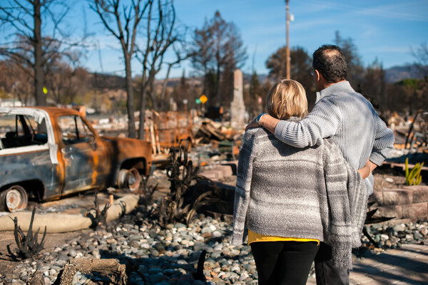 Homeowners inspect the ruined remains of their property after a fire. Rubble, a chimney, and a rusted truck are all that's left.