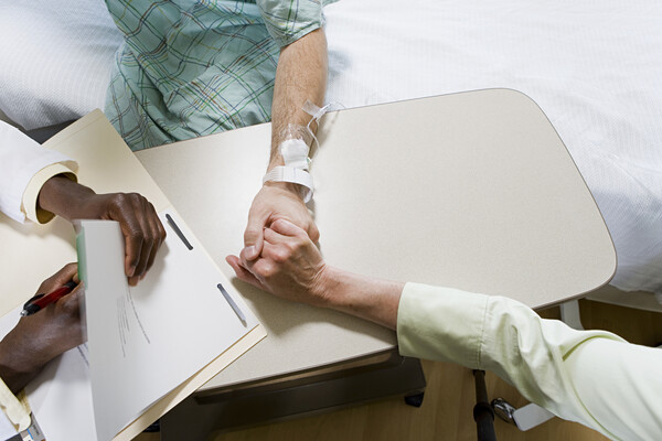A cancer patient holding hands with a relative in consult with a doctor in a hospital bed.
