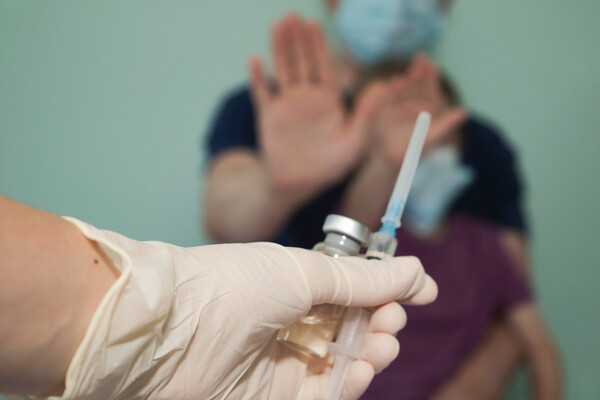 A person with a child holds up a hand to stop a person approaching with a vaccine vial and syringe.