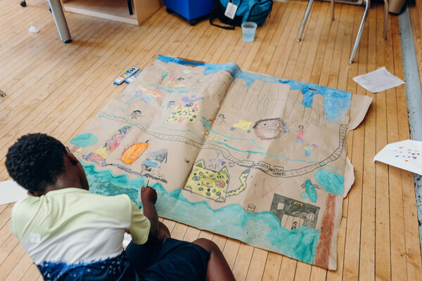 An elementary school student works on a large drawing on the floor of a classroom.