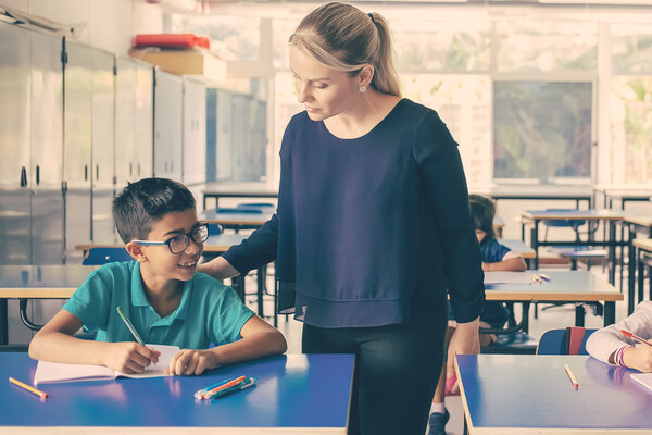 A teacher listens to a young student at their desk.