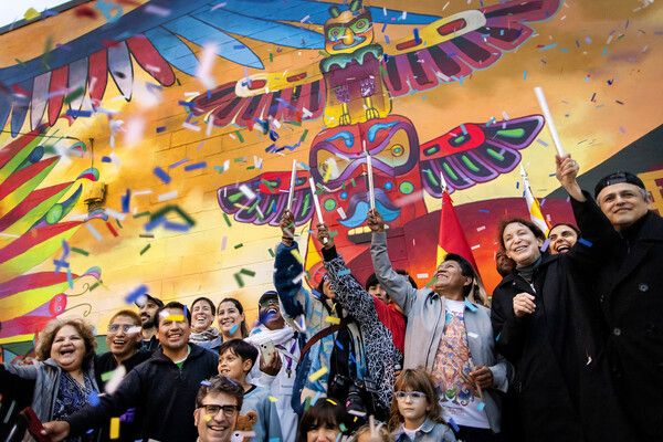 A group of people gather in front of a colorful mural depicting a series of stylized birds. Confetti rains.