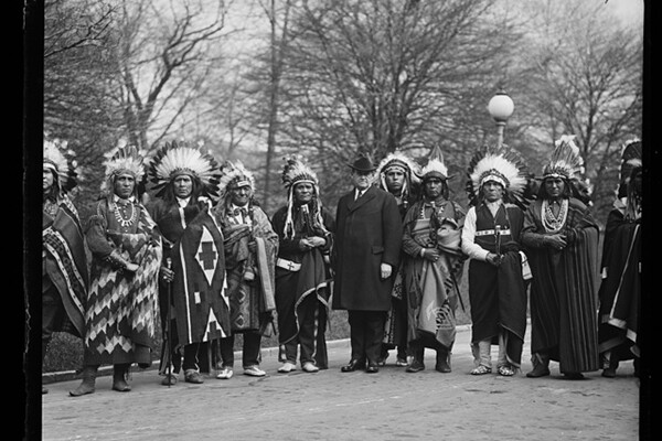 A group of Native Americans standing in a row. The trees behind are bare; it looks to be cold. 