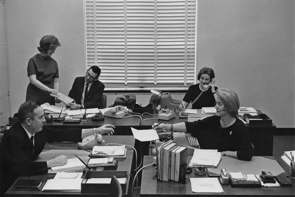Two men and three women sit at desks with typwriters, rotary dial phones and desk calendars in an office in the 1950s.