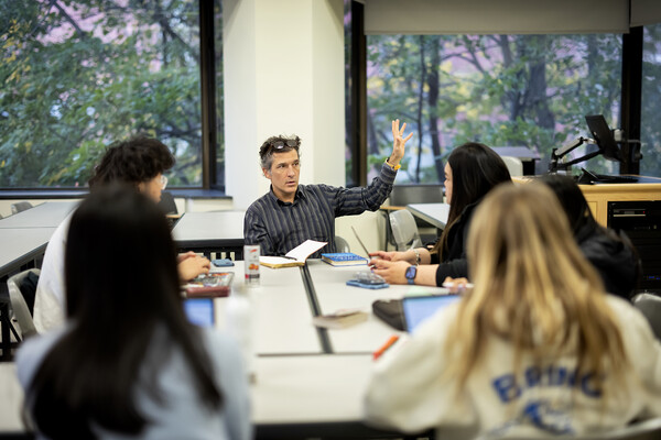 Tyson Smith gestures at the head of a table full of students, in front of windows showing trees.