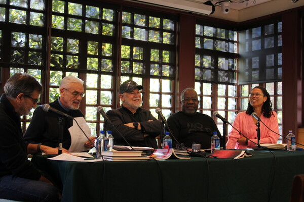 five people sitting at a table with microphones and books in front of a wall of windows