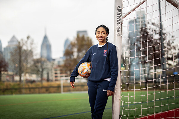 Ginger Fontenot holds a soccer ball while leaning against a goal post at Penn Park, with the Philly skyline in the background.