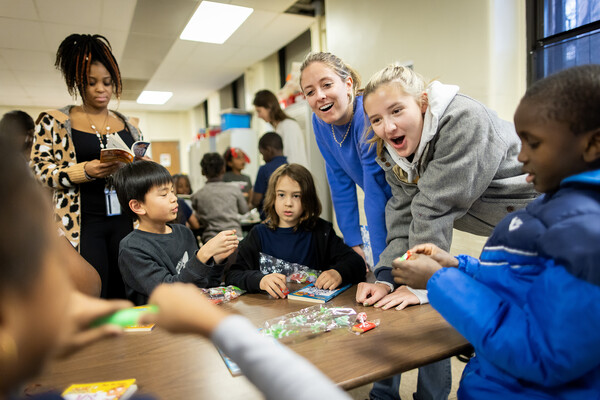 Two college students lean over a table to talk with elementary school students