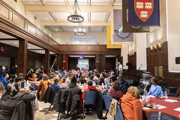 Students sit at tables to eat breakfast in a large room with wooden panelling, chandeliers, and university flags. 