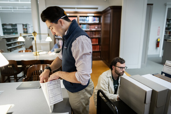 Standing, Joseph Wilbur leaves through documents in a folder; Sam Herrmann crouches near by