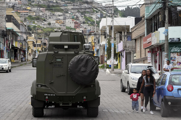 A military vehicle drives through a hilly residential neighborhood in Quito, Ecuador. Two women, one holding hands with a young child, walk alongside on the street. 